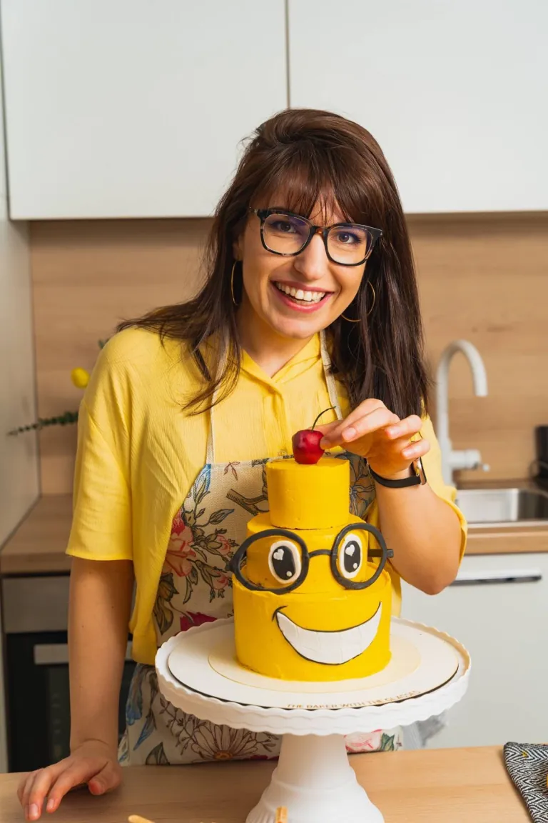Polina smiling in her kitchen, placing a cherry on a two-tier yellow smiley-face cake